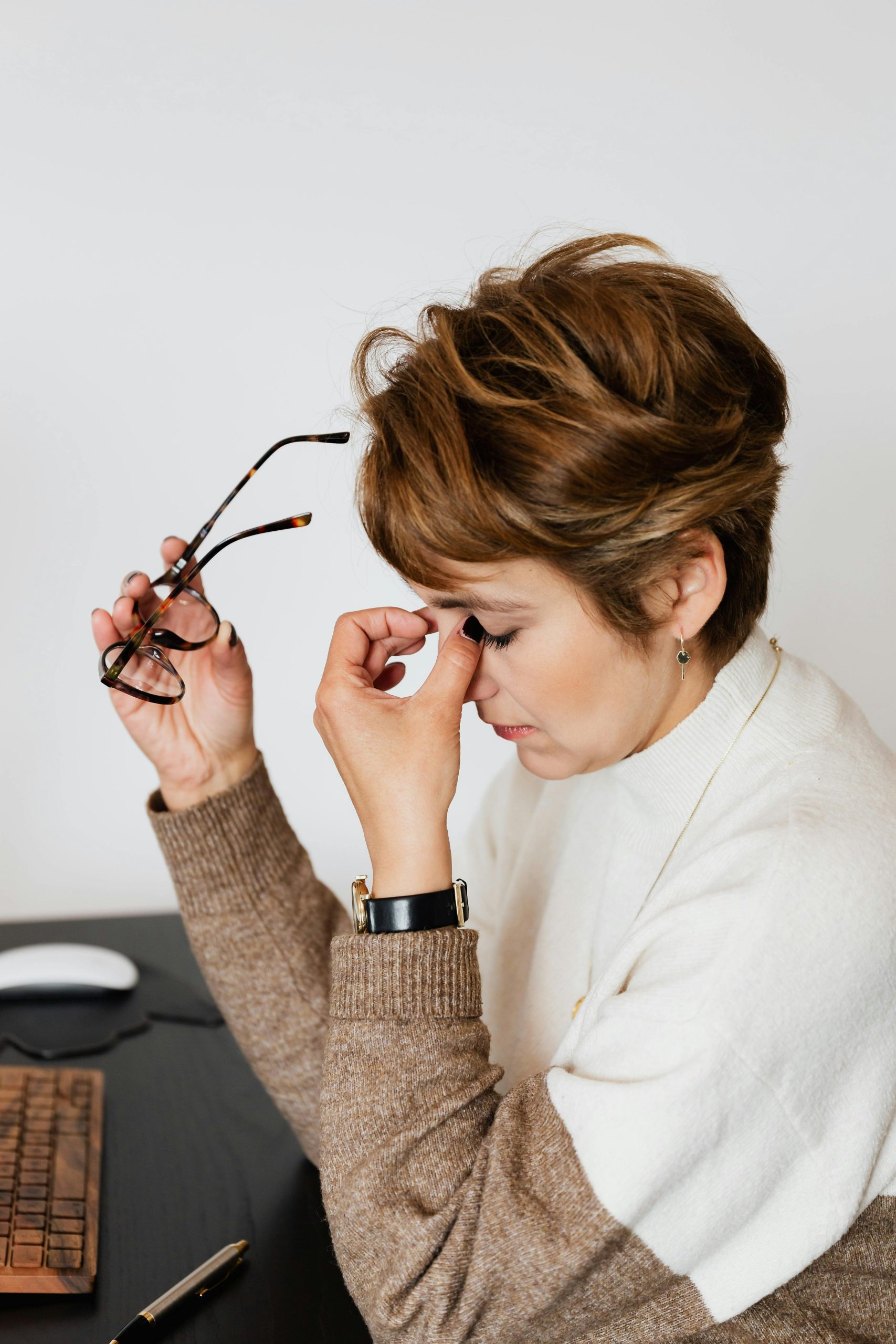 An adult woman feeling stressed and exhausted, holding her eyeglasses at her work desk.